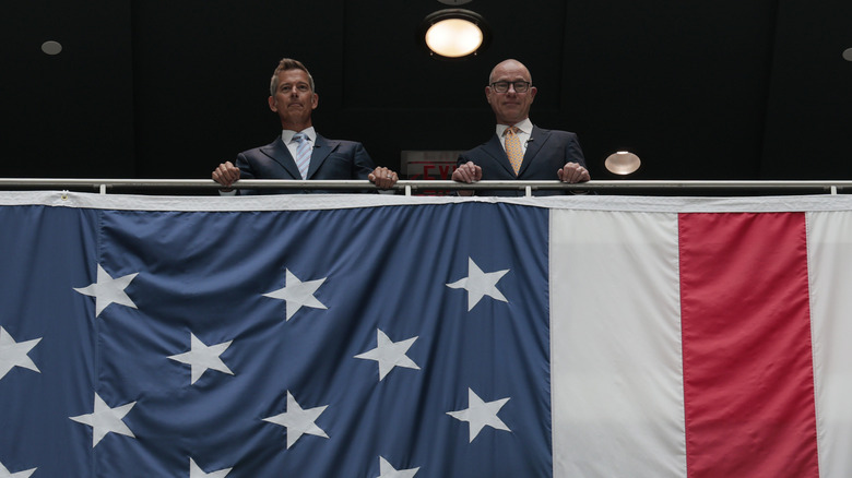Transportation Secretary Sean Duffy looks down over an American flag hanging as a banner from a balcony like something out of the Hunger Games.
